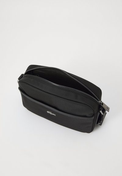 Sac en nylon noir avec une surface texturée, fermeture éclair, poche avant et détails en matériel argenté. Présente un logo de marque à l'avant.