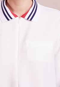 Camicia bianca con colletto a zip, caratterizzata da un colletto a righe in blu marino e rosso. Include una piccola tasca sul petto con contorno cucito.