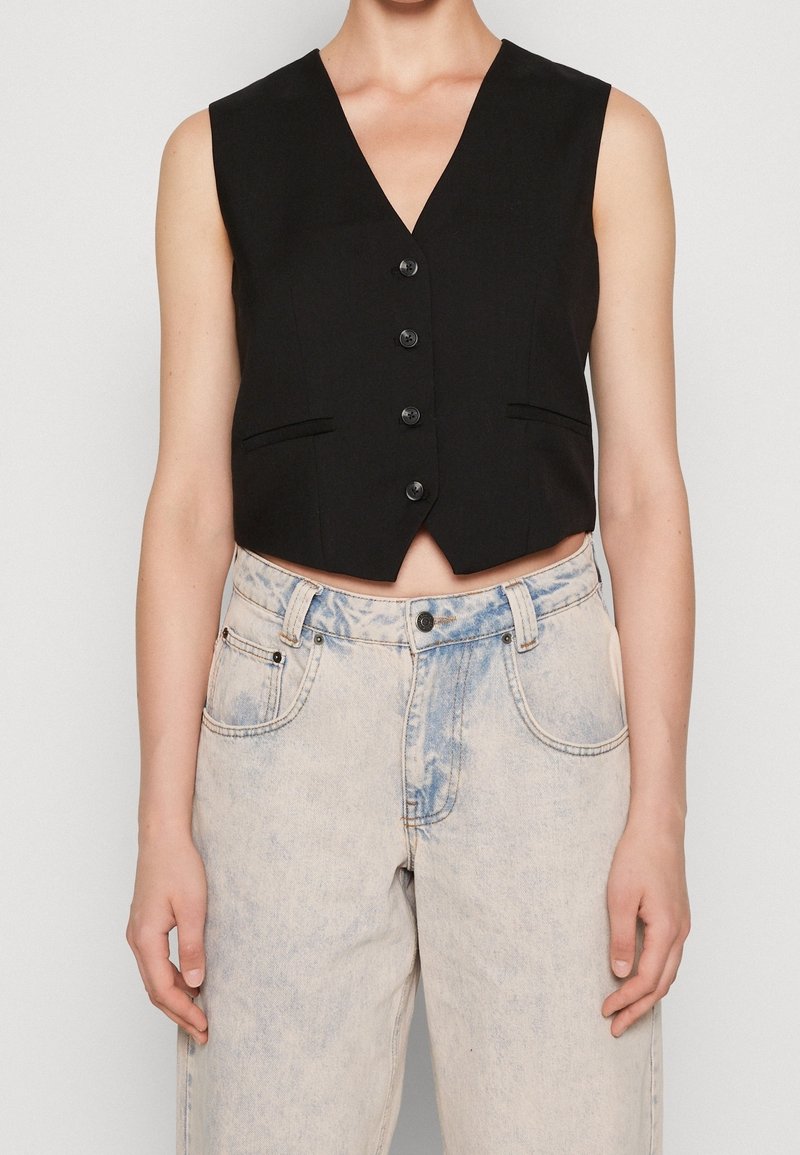 Gilet noir sans manches boutonné associé à un jean en denim clair délavé à l'acide taille haute, porté par une personne debout.