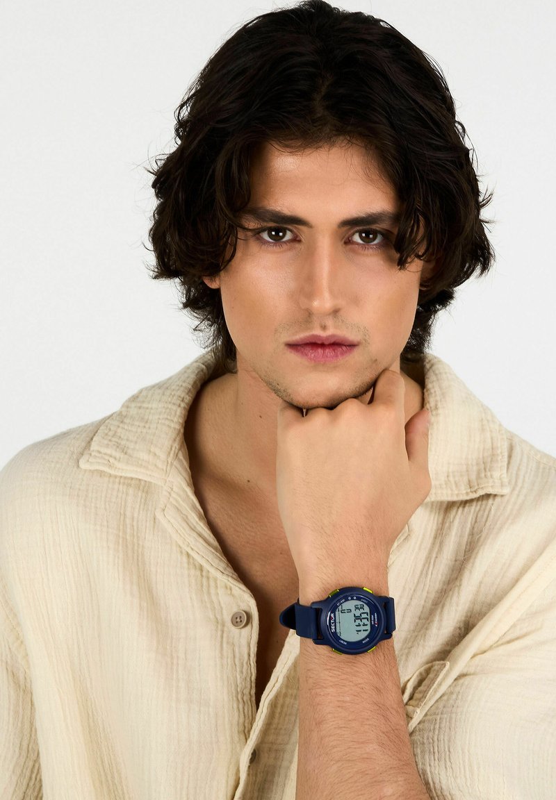 Orologio digitale da polso con cassa tonda blu navy e accenti verdi, dotato di un display LCD chiaro e di un cinturino flessibile. Modello che indossa una camicia chiara.