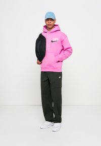 Felpa rosa con logo nero, pantaloni cargo verdi, cappellino azzurro chiaro e in mano una borsa nera. Sneakers bianche offrono un contrasto.