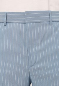 Vaaleansinisiä raidallisia housuja, joissa on litteä vyötärö, sileä pinta ja ohuet valkoiset pystyradat. Yksityiskohtainen ommel taskujen lähellä.