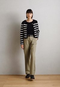 Cardigan a righe bianco e nero sopra una maglietta nera, abbinato a pantaloni khaki e scarpe nere, in piedi su un pavimento in legno.