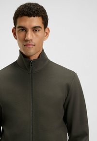 Donkergroene ritsjack met hoge kraag, textuurstof en contrasterende stiksels. Heeft een getailleerd ontwerp en een subtiele glans.