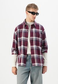 Rød og ternet skjorte med hvide og blå detaljer, som har en lomme og oprullede ærmer, lagd over en cremefarvet sweater og blå jeans.
