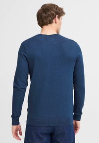 Pulover tricotat bleumarin, cu mâneci lungi, manșete și tiv cu detalii în relief. Materialul pare moale, cu un finisaj texturat. Are un mic etichetă cu logo pe lateral.
