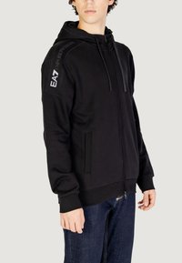 Felpa con zip nera realizzata in tessuto morbido, con cappuccio, chiusura a zip frontale, tasche laterali e logo EA7 sulla spalla.