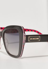 Black oversized sunglasses with gradient lenses, transparent edges, and red heart-patterned temple tips, featuring a gold logo plate.