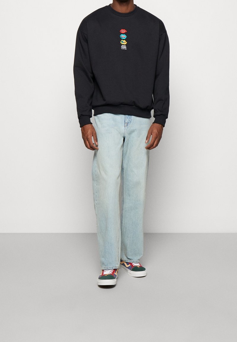 Homme portant un sweat-shirt noir avec un motif de lèvres colorées, un jean bleu clair et des baskets multicolores, debout devant un fond uni.
