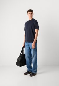 Marine crew-neck t-skjorte, løs passform, sammen med lyseblå rette jeans. Svart duffelbag holdt med en enkelt stropp. Mørke sko.