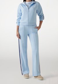 Ljusblå zip-up-träningsdräkt i mjukt tyg. Jackan har en hög krage och en logotyp. Byxorna har sidostrippar och fickor, med en lös passform.