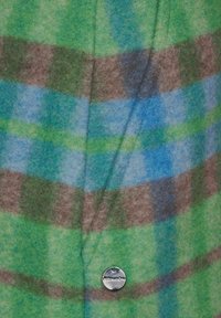 Kangas ruudullista vihreätä ja sinistä väriä, joka on pörröinen; siinä on ruskea ja vaaleanvihreä raita; mukana on pyöreä hopeinen taittu, jossa on tekstiä.