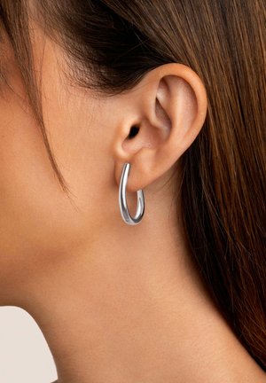 Silberner Creolenohrring, der an einem durchstochenen Ohr präsentiert wird, mit einer glatten, reflektierenden Oberfläche und einem zeitgenössischen, länglichen Design.