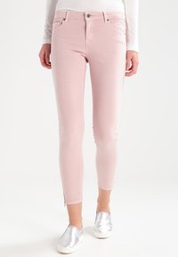 Een vrouw in lichtroze slim-fit broek, gecombineerd met zilveren instap schoenen en een witte ribbeltrui met lange mouwen, staande voor een witte achtergrond.