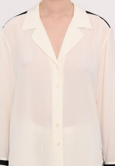 Camicia button-up leggera beige chiaro con colletto, caratterizzata da dettagli neri lungo le spalle e una linea dritta. Texture morbida e liscia.