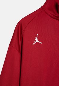 Sarkans sporta žakets ar augstu apkakli, uz krūtīm ar balto izšūto Jumpman logo. Gluda, viegla auduma sastāvs ar teksturētu apdari.