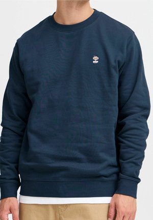 Homme portant un sweat-shirt bleu marine à col rond avec un petit patch brodé de dessert sur la poitrine, un pantalon beige et un sous-shirt blanc visible.