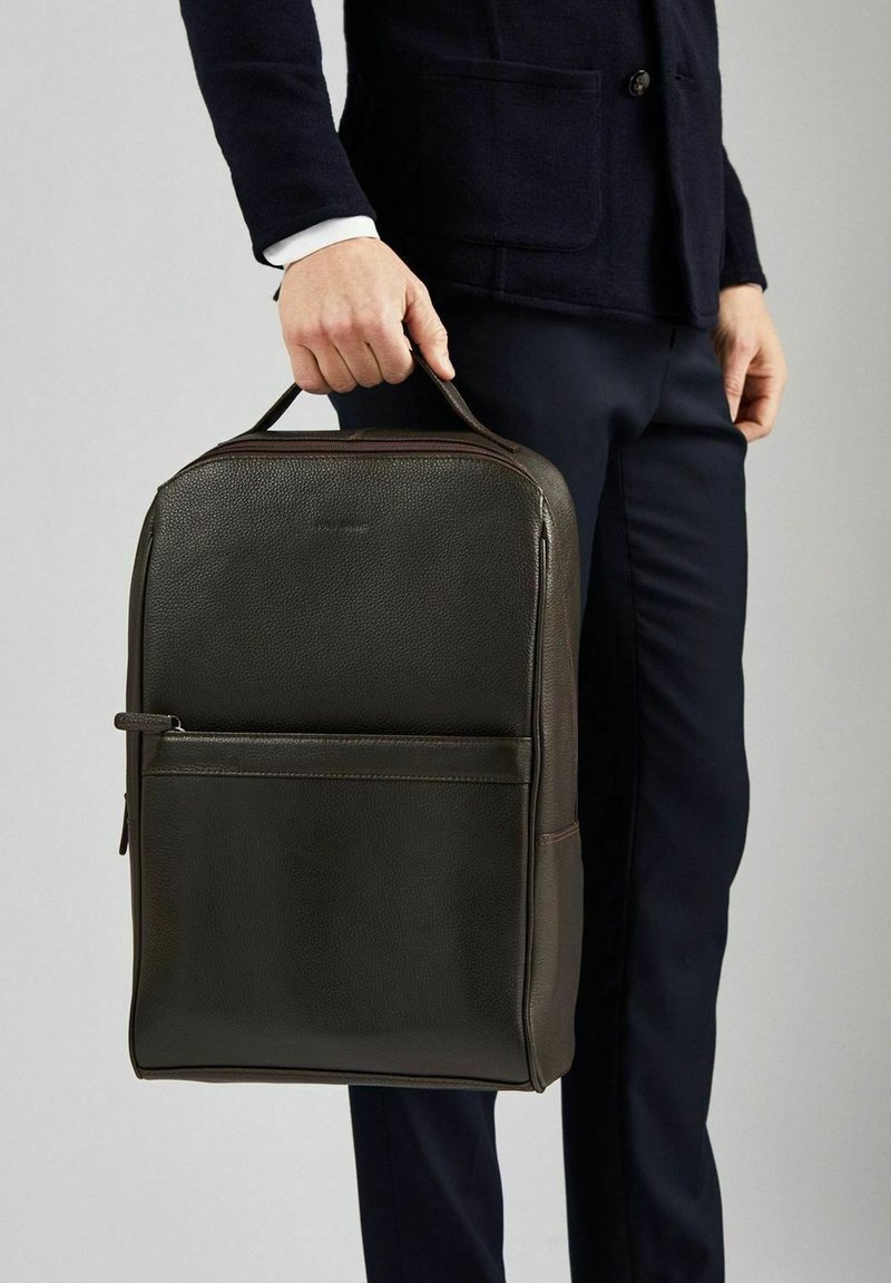 Mörkbrun läderryggsäck med texturerad yta, utrustad med ett handtag på toppen, framfickficka med dragkedja och minimalistisk design.