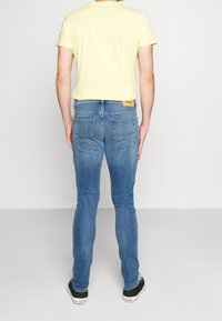 Gul t-shirt med korta ärmar och figursydd modell. Ljusblå tajta jeans med två bakfickor, diskret blekning och varumärkesetikett på midjebandet. Svarta sneakers.