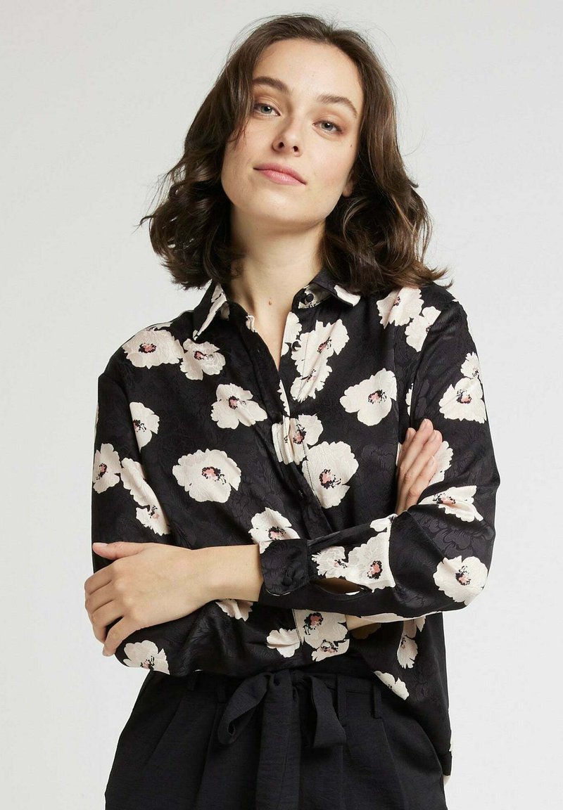 Chemise noire avec motif floral blanc, design à boutons, manches longues et col. Texture lisse avec un léger éclat et poignets avec détails de boutons.