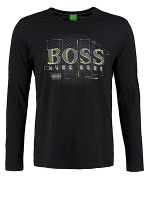 Mann trägt schwarze Hose, schwarzes T-Shirt mit "BOSS"-Logo, schwarzen Cardigan mit gelber Einfassung und schwarze Sneakers, steht mit einer Hand in der Tasche.