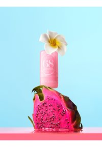 Różowa tuba balsamu perfumowanego z białym kwiatem na górze, zrównoważona nad żywą różową połówką owocu smoka na niebieskim tle.