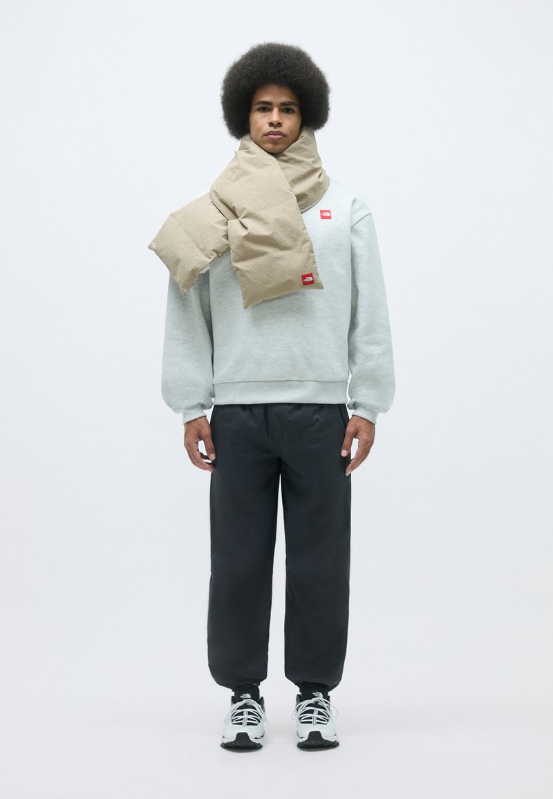 Helle graue Sweatshirt mit rotem Logo, schwarze Sporthosen und ein beiger Schal, der um den Hals drapiert ist; Model steht vor einem weißen Hintergrund.