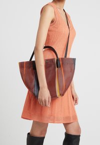 Transparante net tote bag met een bruine basis, voorzien van meerkleurige strepen en zwarte leren handvatten. Draagbaar over een kort oranje jurk.
