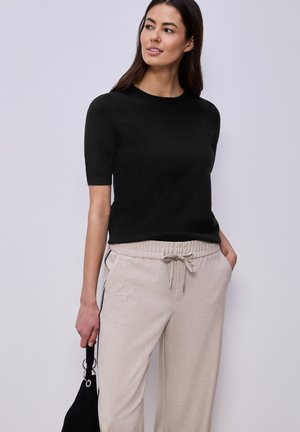 Frau trägt ein schwarzes kurzärmeliges Oberteil und beige Hose mit Kordelzug, hält eine kleine schwarze Handtasche und steht vor einem hellgrauen Hintergrund.