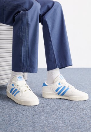 Witte sneakers met blauwe accenten, voorzien van drie blauwe strepen en een gestreepte zool, gecombineerd met grijze sokken en marineblauwe broek.