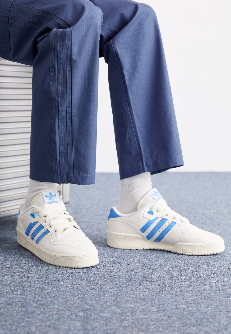 Vita sneakers med blå accenter, med tre blå ränder och en texturerad sula, i kombination med grå strumpor och marinblå byxor.