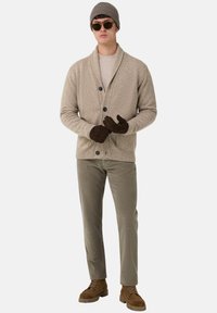 Man med beige kofta, ljus tröja, grå mössa, solglasögon, bruna vantar, taupefärgade byxor och bruna stövlar som står mot vit bakgrund.