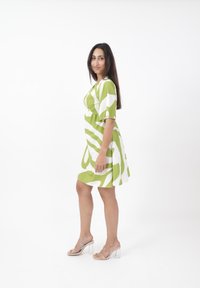 Grün-weiß gemustertes kurzes Kleid mit tailliertem Oberteil und ausgestellt rock. Das Modell trägt transparente High-Heelsandalen. Einfarbiger weißer Hintergrund.