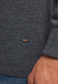 Grauer gestrickter Pullover mit geripptem Saum und Bündchen. Verfügt über ein kleines braunes Akzentetikett nahe dem unteren Rand. Das Material wirkt weich und strukturiert.