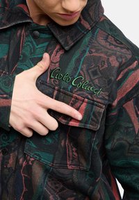 Lange mouw shirt in zwart, rood en groene stof met een gestructureerd patroon. Voorzien van een borstzak en een geborduurd logo in groene draad.
