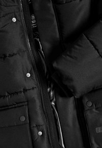 Sort pufferjakke med en blank inderside, der har lynlås lukning, trykknap og quiltet yderside. Tekstureret stof med to lommer.