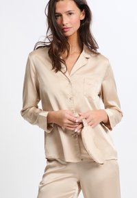 Satin-beiges Pyjama-Set mit einem knopfbaren Hemd mit einer Tasche und langen Ärmeln, kombiniert mit passenden Hosen und einer Augenmaske.