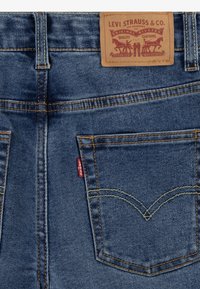 Blue denim jeans featuring a leather label with embossed graphics, orange stitching, and a signature red tab on the back pocket.