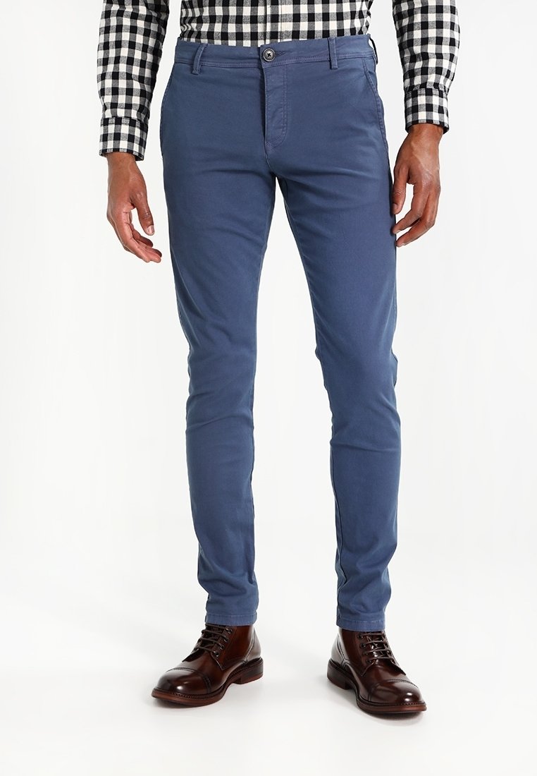 Man med blå slim-fit byxor, bruna läderskor och svartvit rutmönstrad långärmad skjorta, stående mot vit bakgrund.