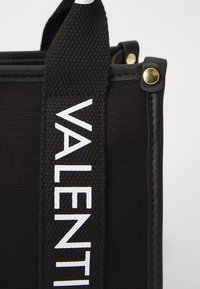 Black fabric tote bag with a textured finish. Features a prominent white logo, gold hardware accents, and sturdy top handles.