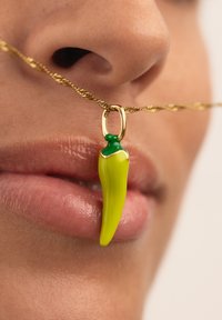 Ciondolo a forma di peperoncino giallo su una catena dorata attorcigliata, con un accento verde sulla parte superiore. Ambientato su una pelle liscia con una sottile texture delle labbra.
