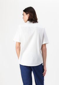 Camicetta bianca a maniche corte con una vestibilità rilassata e orlo curvo. Il materiale sembra leggero e traspirante, adatto per un abbigliamento casual.