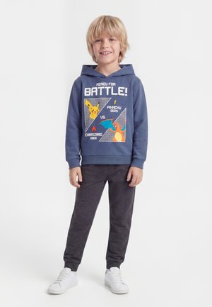 Lyshåret dreng smilende, iført en blå hættetrøje med Pikachu- og Charizard-grafik, mørke bukser og hvide sneakers, stående på en hvid baggrund.