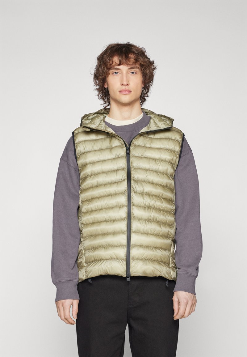Ljus olivgrön puffer-vest med ett quiltat mönster, hel dragkedja och huva; bärs över en grå sweatshirt med svarta detaljer och fickor.