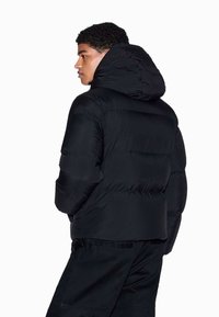 Schwarze Daunenjacke mit Kapuze, mit gesteppter Optik und elastischen Bündchen. Getragen von einer Person, die von hinten blickt, seitlich gesehen.