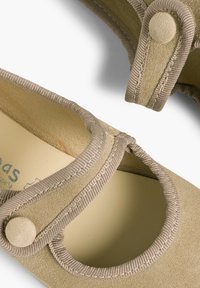 Zapato de ante beige con puntera redondeada, correa elástica con cierre de botón y forro interior suave. Cortes curvados para un detalle adicional.