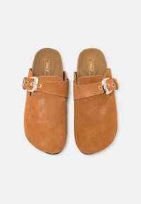 Sabots en suède marron à enfiler avec une large sangle et un détail en boucle argentée. Surface texturée et design à bout rond. Doublure intérieure douce.