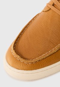 Zapato de cuero marrón claro con una textura suave, que presenta un borde cosido prominente y una suela de goma blanca. Un diseño limpio y minimalista acentúa su forma.