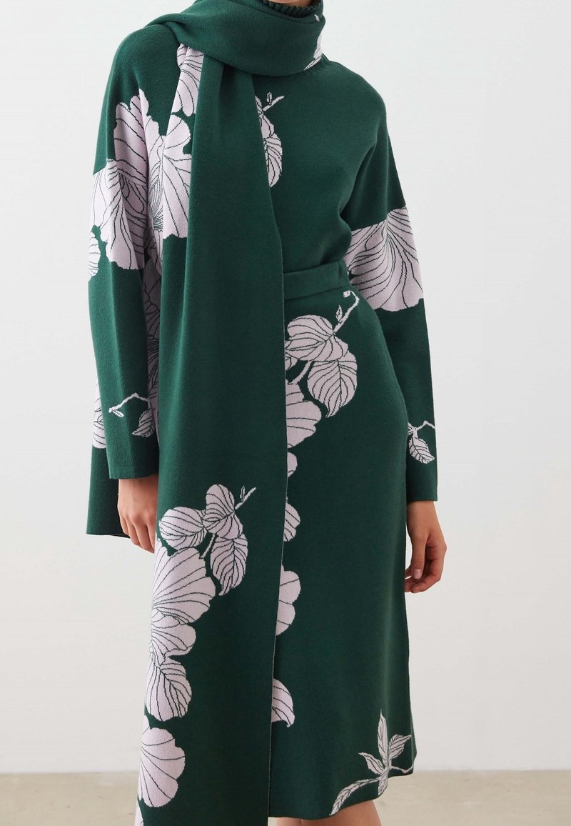 Robe à manches longues vert foncé et foulard avec de grands motifs floraux de feuilles rose pâle, portée par une personne debout devant un fond clair uni.