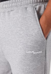 Sive pamučne trenirke s elastičnom strukom i bočnim džepovima, s bijelim logotipom "LOVE SCARS" na prednjem dijelu nogavice.